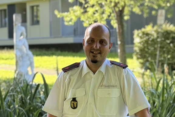 Eine männliche Person steht auf einer Brücke und lächelt in die Kamera. Er hat ein weißes Feuerwehrhemd und schwarze Hose an. 